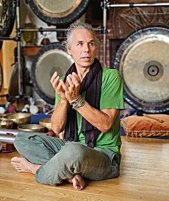 Joseph sitting on the floor with legs crossed, lotus mudra with gongs behind him