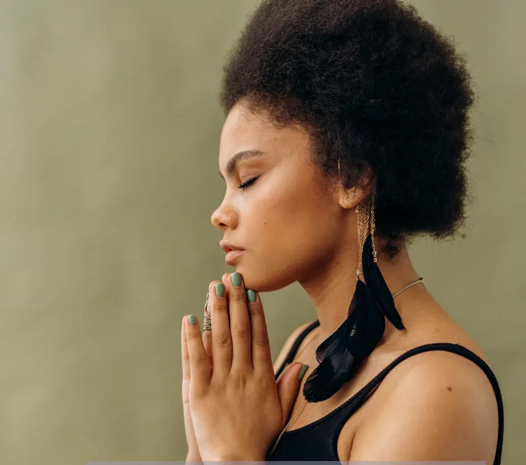 A woman is contemplating healing potential. She is standing with her eyes closed, with prayer hands at heart. She has dark skin, black hair and wearing long black feather earrings that brush her collar bone.