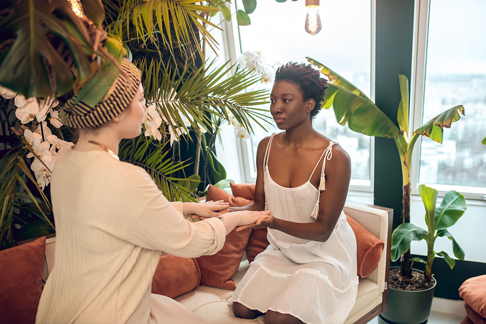 Sangha Healing Holistic Practices - A holistic practitioner with a turban on her head sitting across from her client, wearing white dress. They are touching hands and sharing energy.